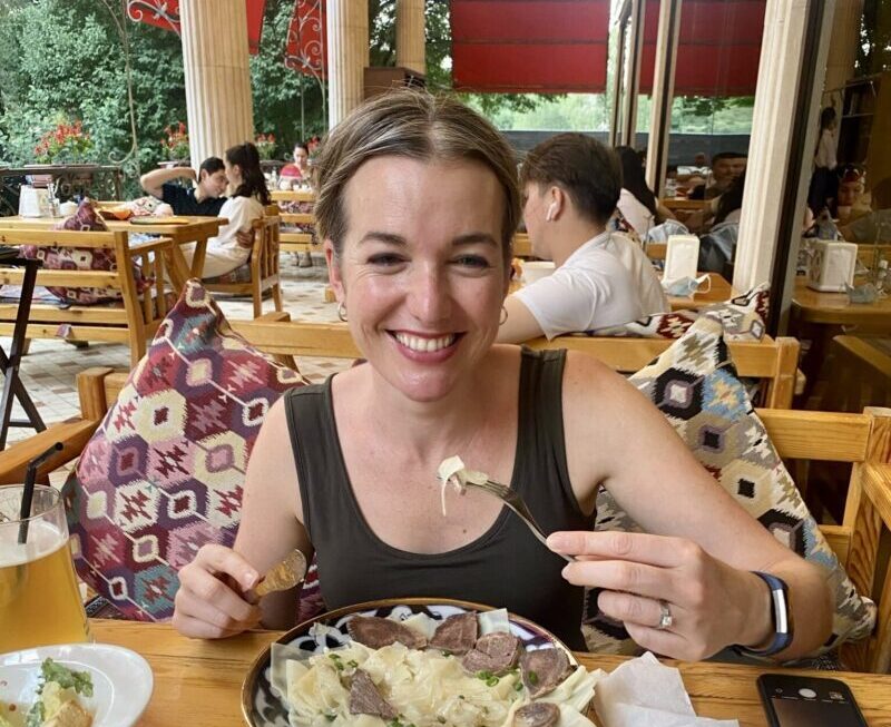 A woman sitting at a table full of food, including Kazakhstan's national dish, Beshbarmal, holds up a forkful and smiles at the camera.