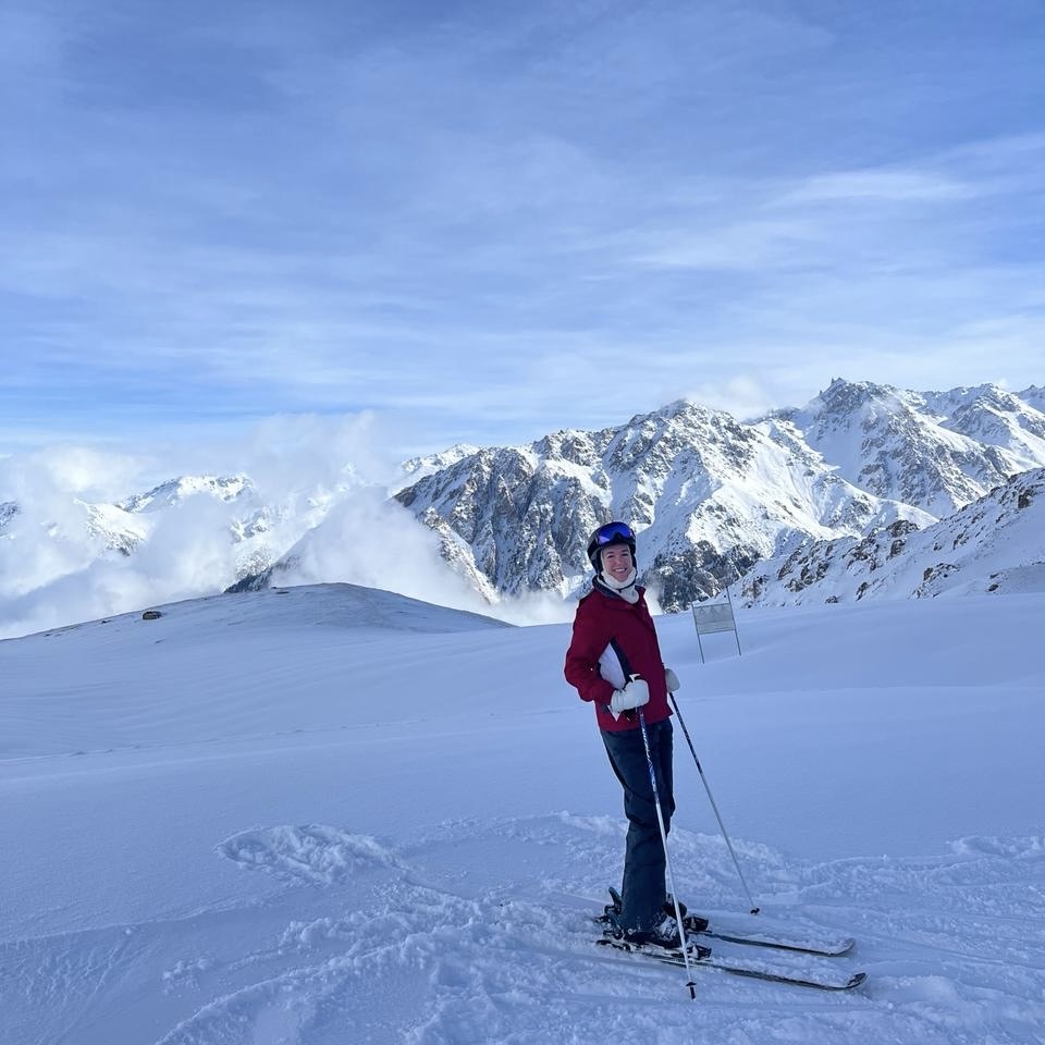 A woman in ski gear stands on a ski slope in front of a large snowy mountain range. Shymbulak is a great choice for lovers of snow sports, as well as lovers of epic mountain scenery and fresh mountain air.