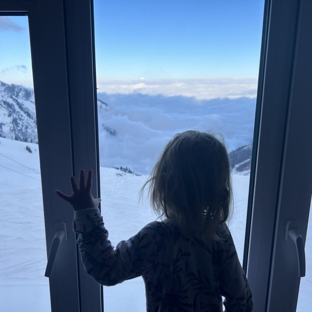 A small child looks out the window from above the clouds at a mountain vista with 