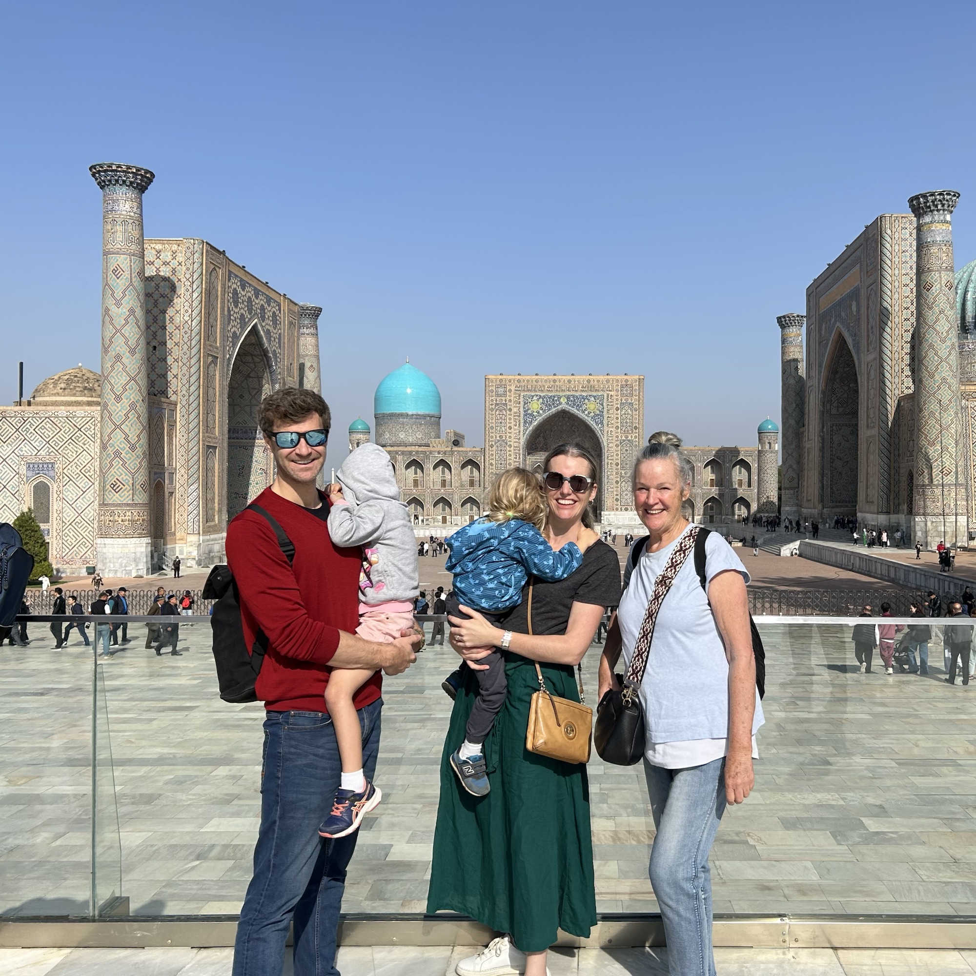 A family with two small children stand in front of Register Square, Uzbekistan, a beautiful and monumental example of muslim architecture with blue mosaics.