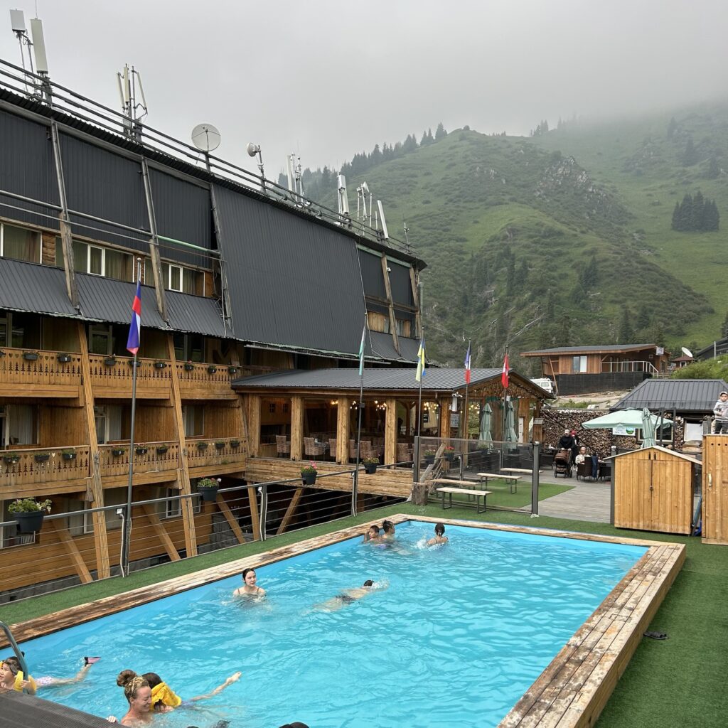 People swim in a pool in front of a hotel considered a great place to stay at Shymbulak Resort, with misty mountains in the background