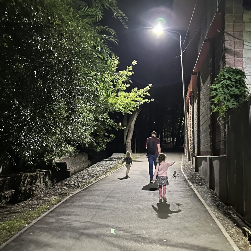 A young family walk up a pathway at night with a bright streetlight above