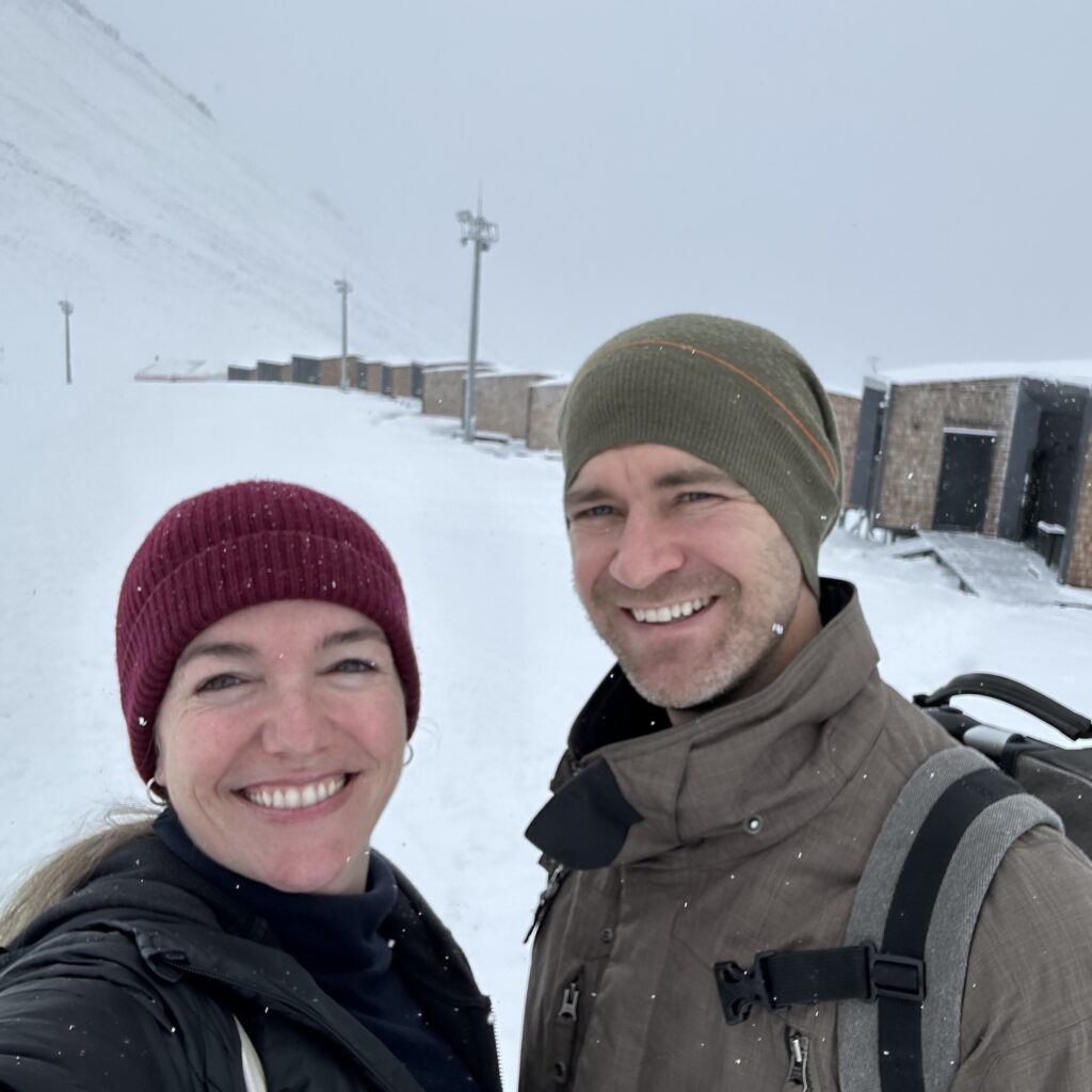 A man and woman stand in the snow in front of Tenir Hotel, a series of cabins in Shymbulak Resort