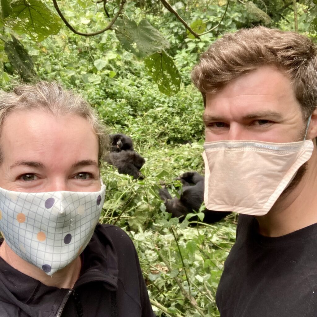 a woman and a man in masks stand in the undegrowth in front of a family of gorillas