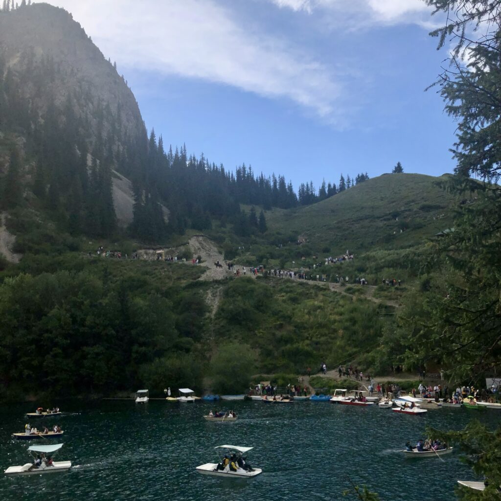 Kolsai Lake in the foreground with many tourists on rowboats and paddle boats on it and the mountain and blue sky behind, with many more tourists heading down towards the lake