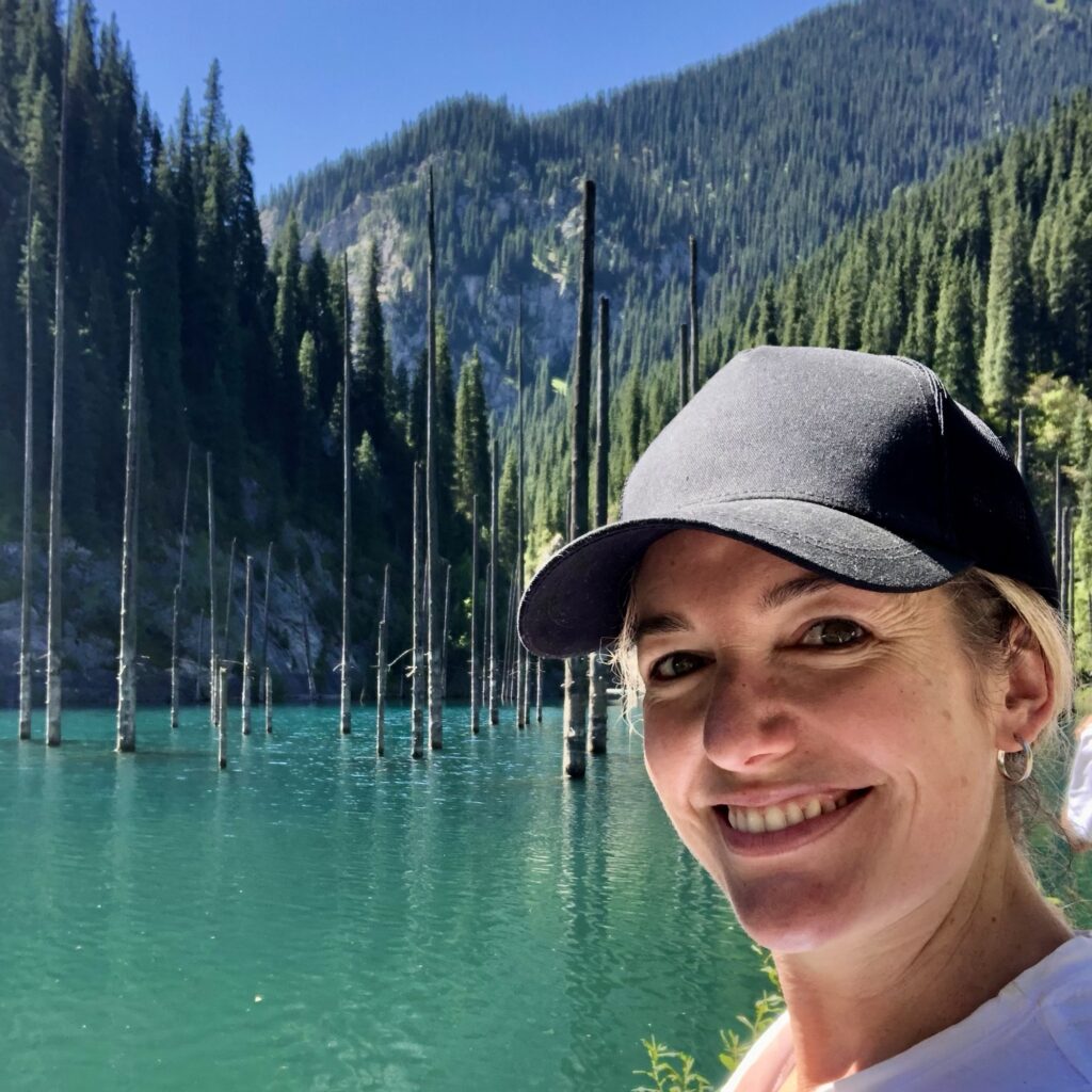 A woman smiles in front of greenish blue Kaindy Lake with trees poking out of it in, 