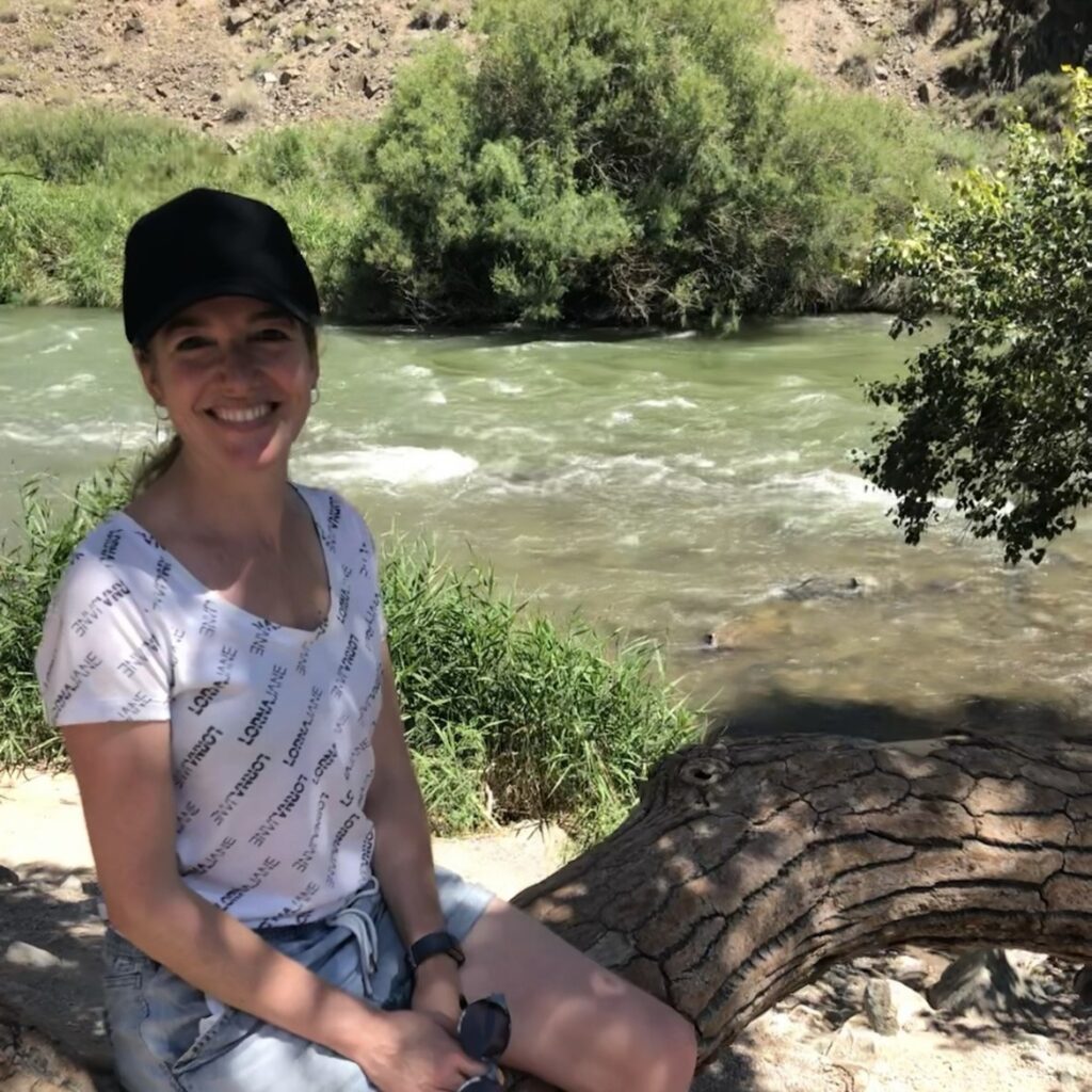 a woman smiles at the camera from the branch of a tree with the very rushy Charyn River behind her 