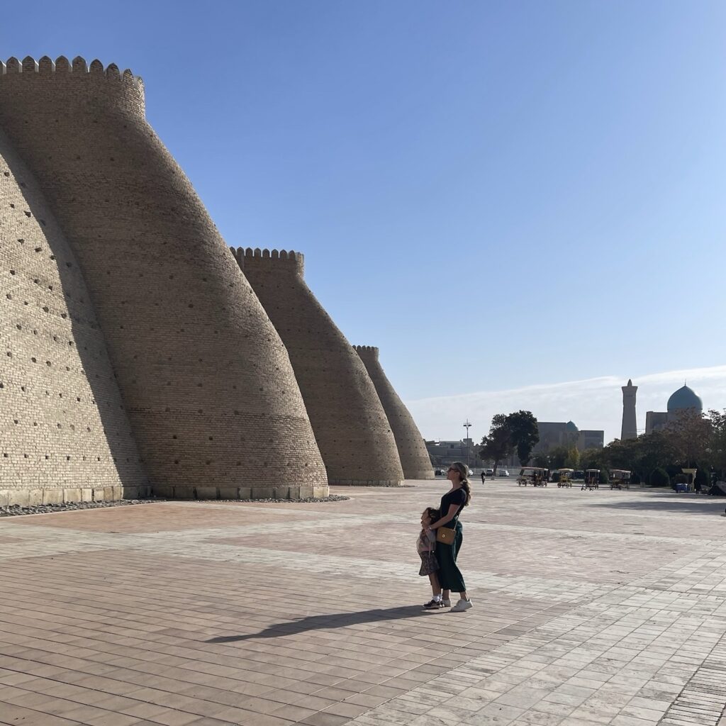 A woman and a child stand facing the imposing rounded walls of the Ark of Bukhara, an impressive ancient fortress in Uzbekistan 