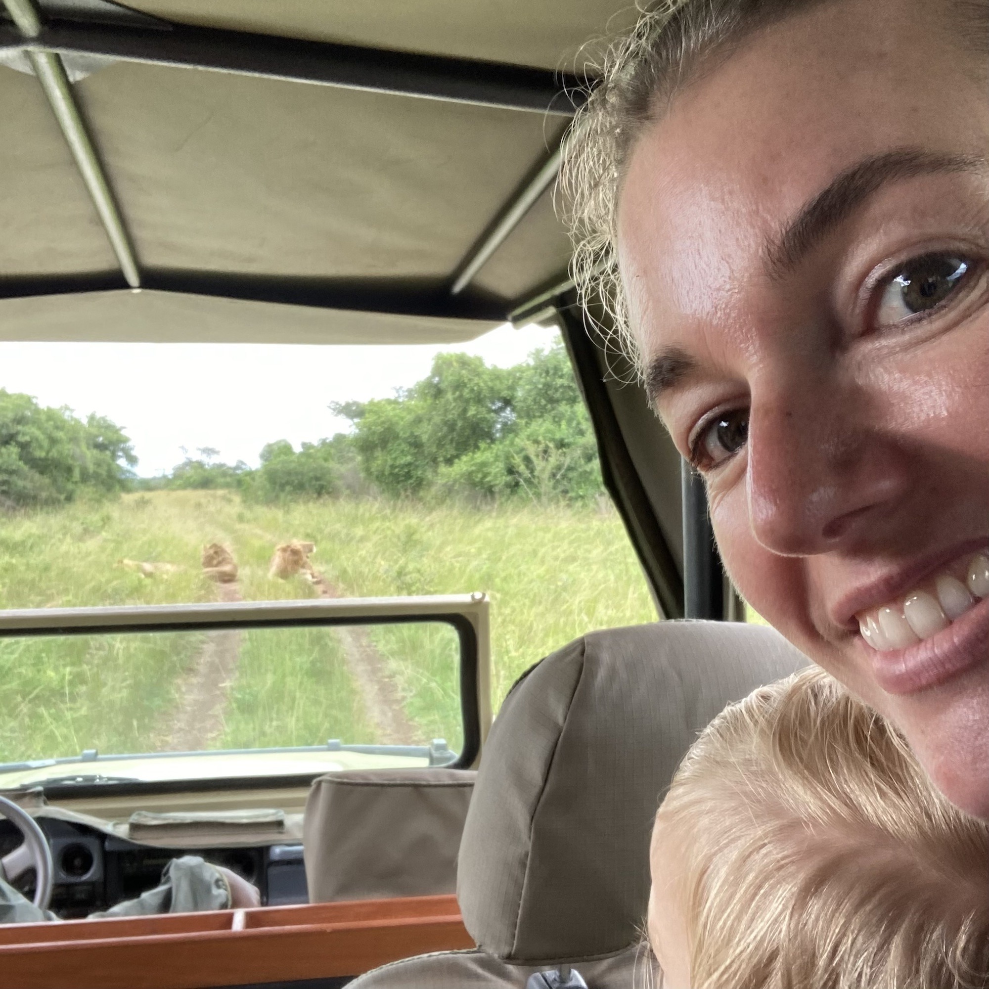 a woman smiles at the camera in the foreground while in the background some of Akagera's lions recline in the sun