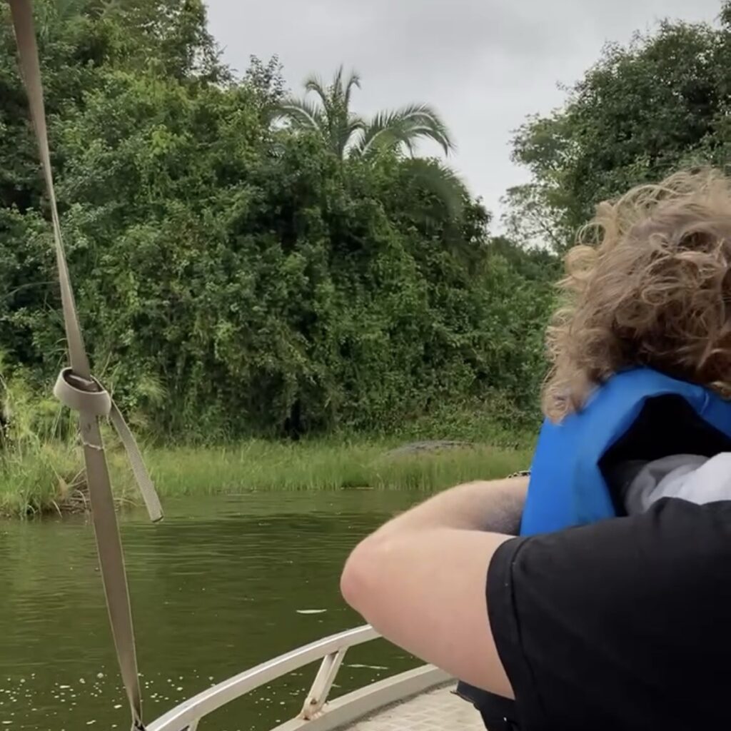 a child holds onto their father as their boat approaches land with a large crocodile laying in the grass. This is in Lake Ihema at Akagera Safari Park