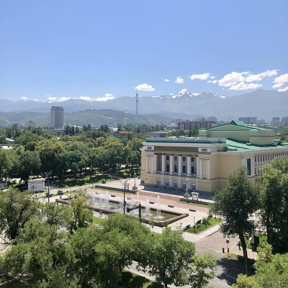 City of Almaty with the Abay Theatre in the foreground, and Kazakhstan Hotel, Communications Tower, and Tier Shan Mountains in the background.