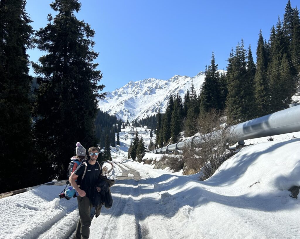 a hiker smiles at the camera with a small child on his back and the snowy road to Big Almaty Lake behind him