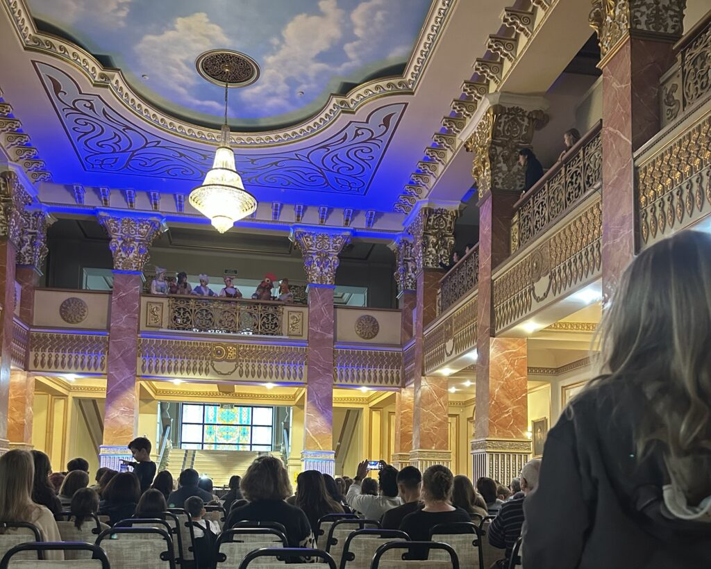 A child looks on in an ornately decorated hall as opera singers perform on the balcony ahead