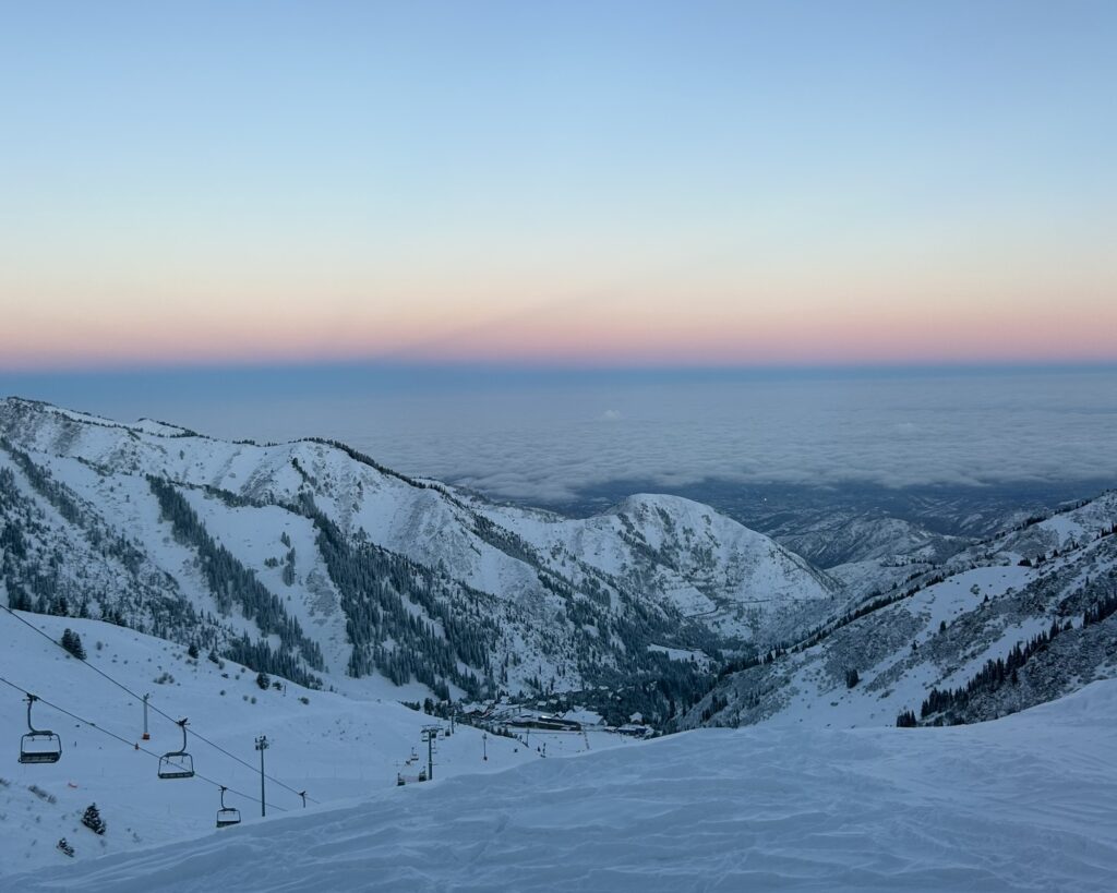 looking down over a ski slope with some small mountains to the sides and a cloud covered city in the background