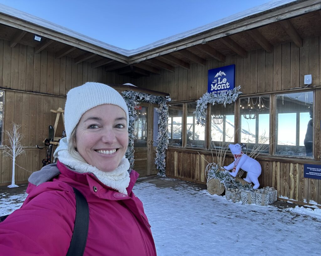 A woman in ski gear stands in front of the snowy entrance to ski lodge Le Mont Hotel