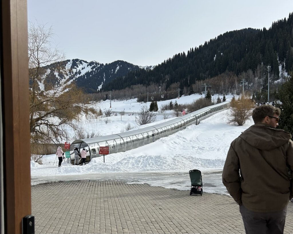 A man stands to the side in the foreground looking out at Ak Bulak Ski resort with the entrance of a magic carpet lift in near distance