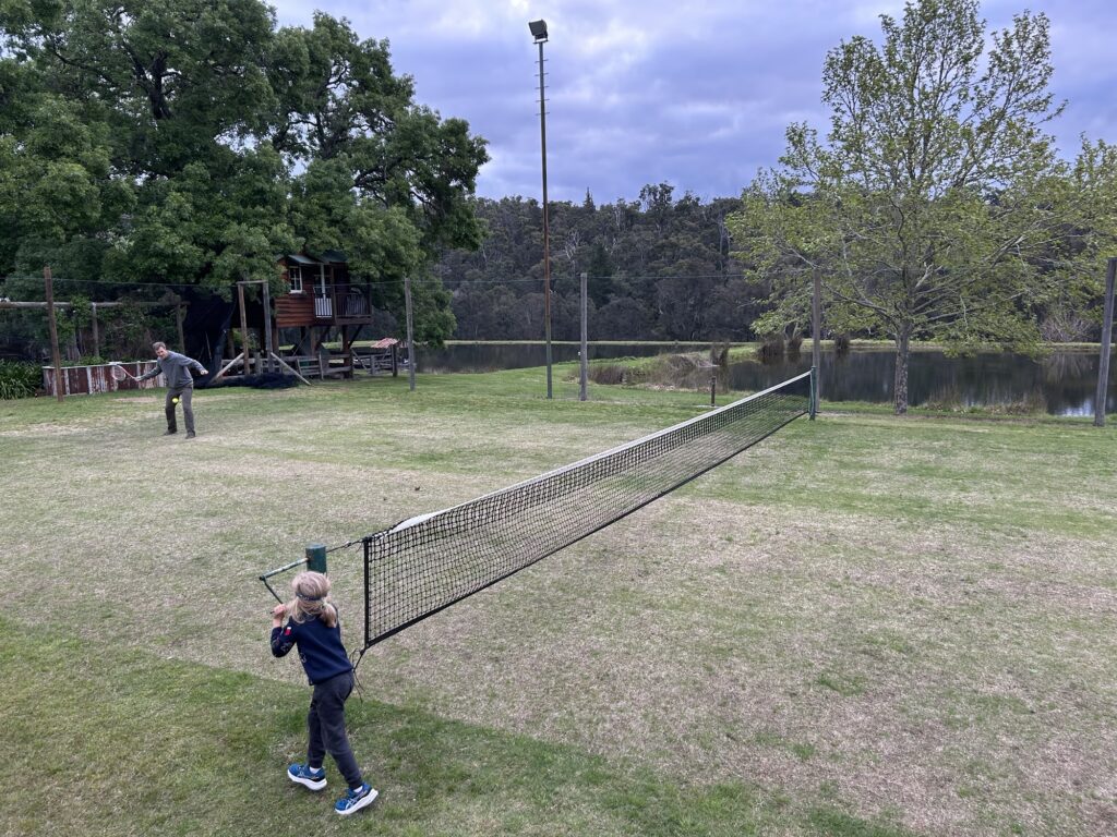 A photo of a man playing tennis on a grass court while a child tightens the net, at Clover Cottage Manjimup