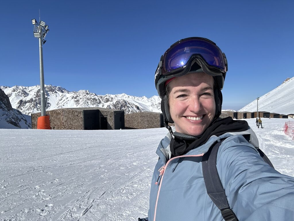 A photo of a woman in ski gear smiling in front of Tenir Eco Hotel on top of Shymbulak Mountain Resort