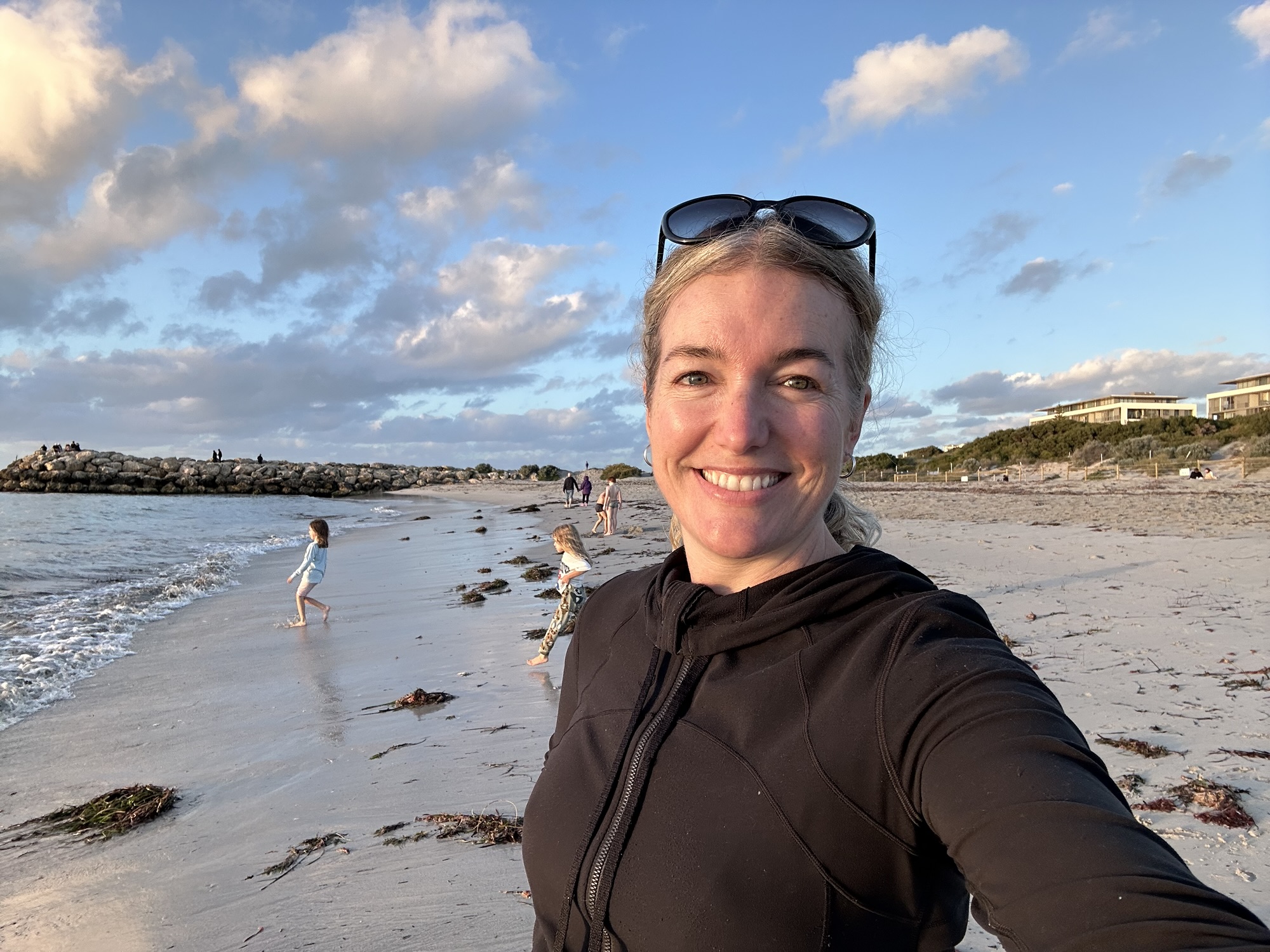 A photo of a woman in the foreground smiling at the camera while children behind her play at the beach