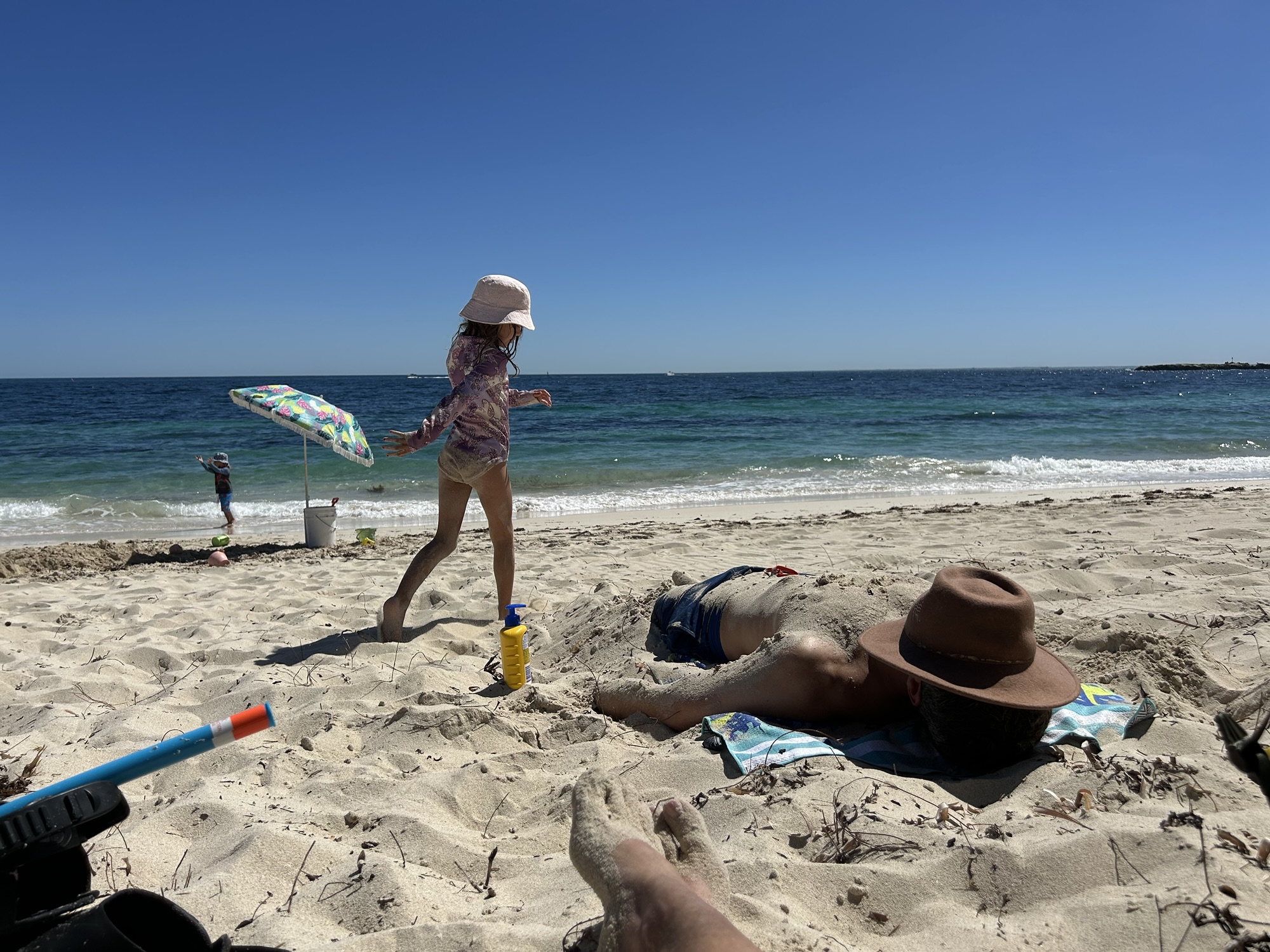 A photo of a girl walking along a beach on Rottnest Island with her father partially buried in the sand near her feet. Rottnest is a great stop for a 7 day family itinerary in Western Australia