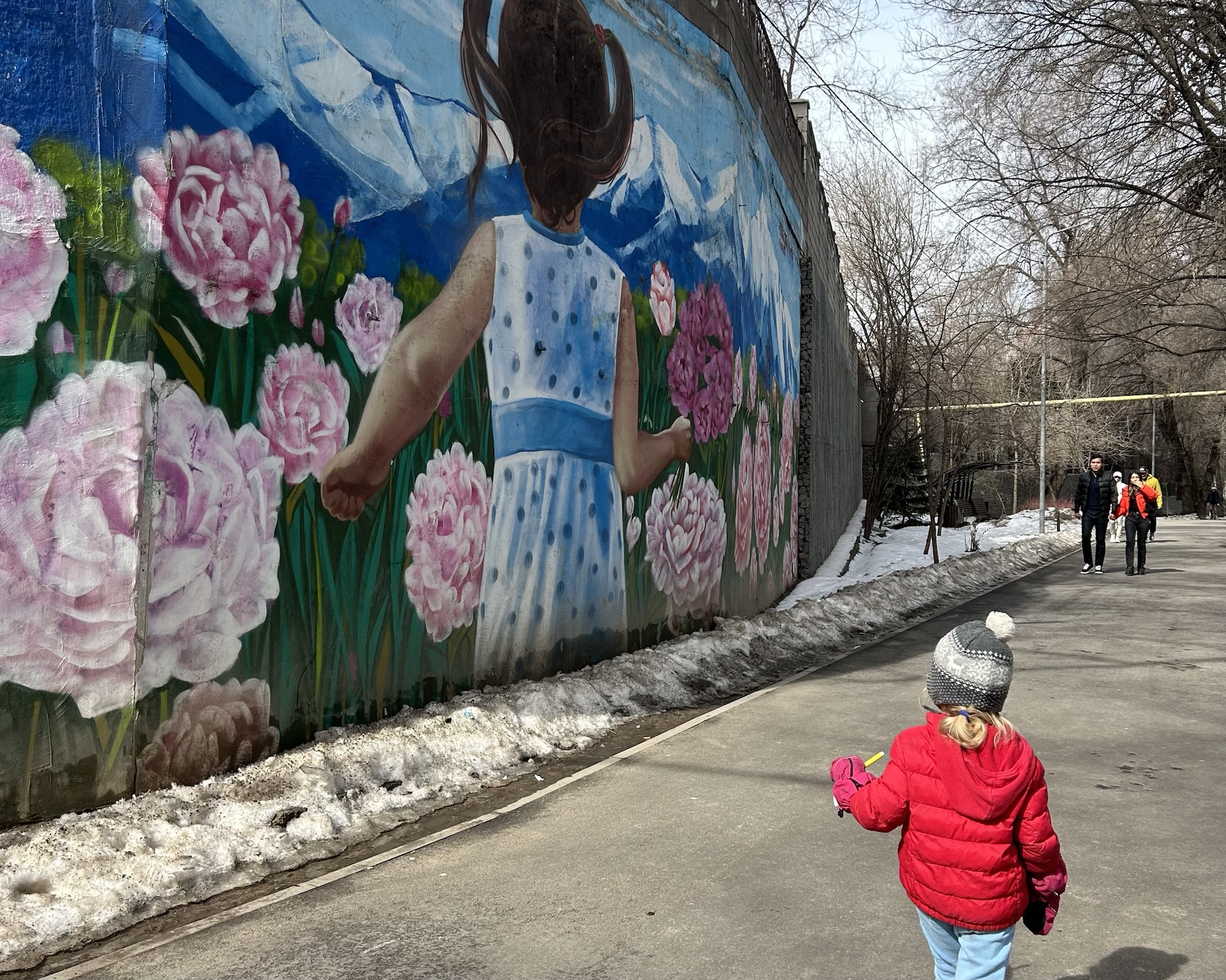 A photo of a small child in the foreground looking back at a mural of a girl surrounded by flowers on the wall of the Terrenkur river path in Almaty, Kazakhstan