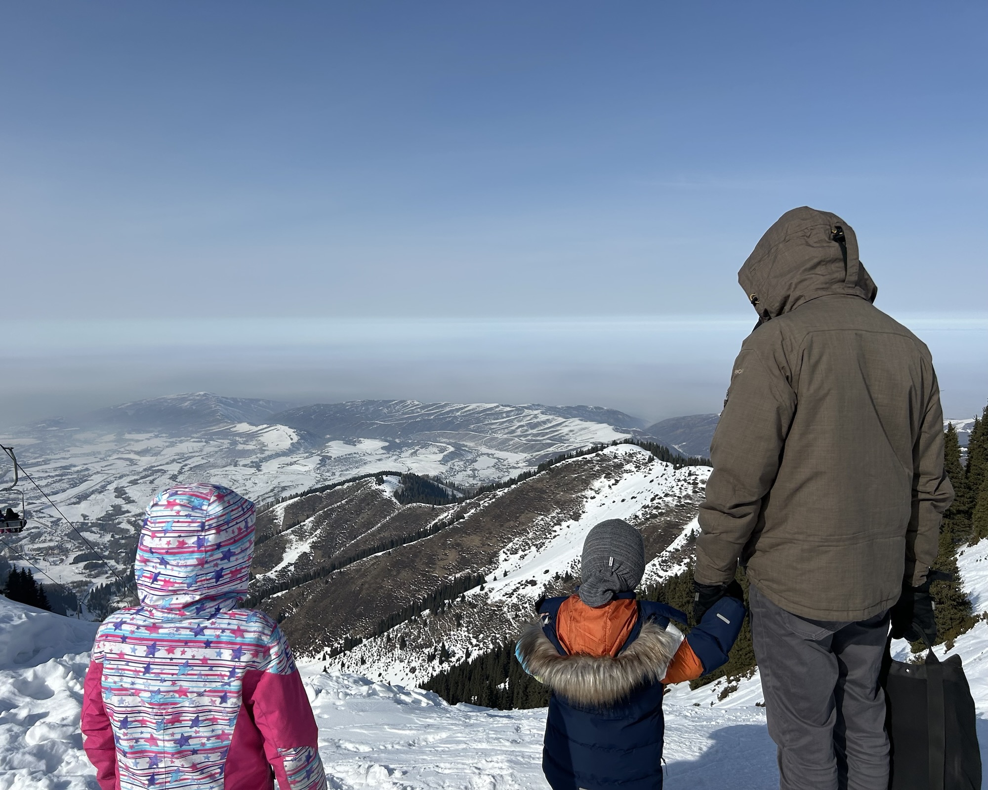 A photo of a man and 2 children in snow gear looking out over some snow covered mountains in Kazakhstan